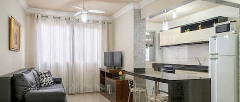A modern kitchen with a central island, a dining area with bar stools, a sofa with a TV, and a ceiling fan.