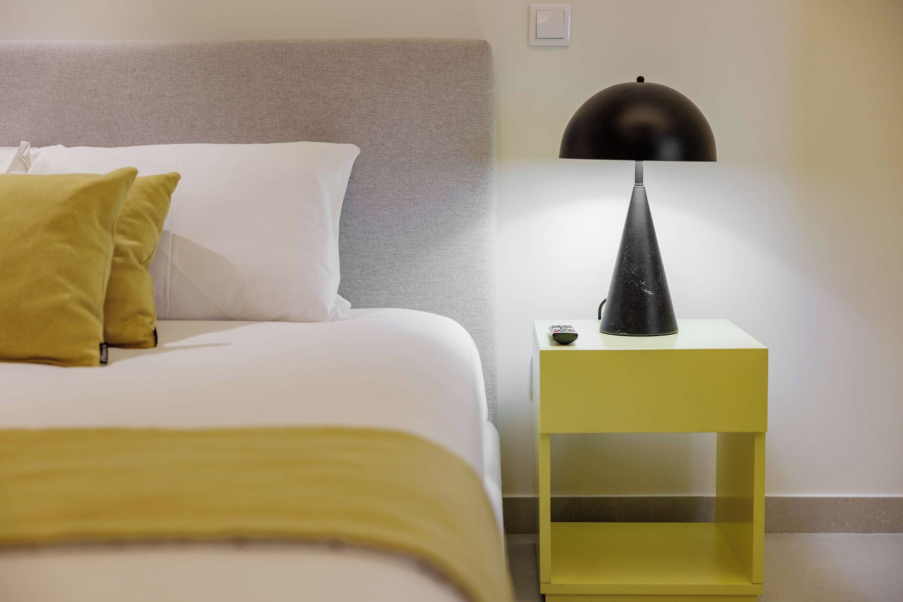 Ein ordentlich gemachtes Bett mit weißen und senffarbenen Kissen, einem grauen Kopfteil und einem gelben Nachttisch mit einer schwarzen Lampe.