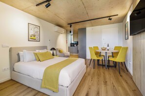 Une chambre d’hôtel moderne avec un lit, un coin repas avec des chaises jaunes, une cuisine en arrière-plan et un téléviseur fixé au mur.