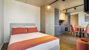 A modern hotel room with a large bed, a wooden accent wall, a kitchenette, and a dining area with red chairs. - Marina da Afurada Apartments - Fernando Pessoa (Vila Nova de Gaia)