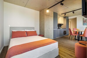 Ein modernes Hotelzimmer mit einem großen Bett, einer Wand mit Holzvertäfelung, einer Küchenzeile und einem Essbereich mit roten Stühlen.