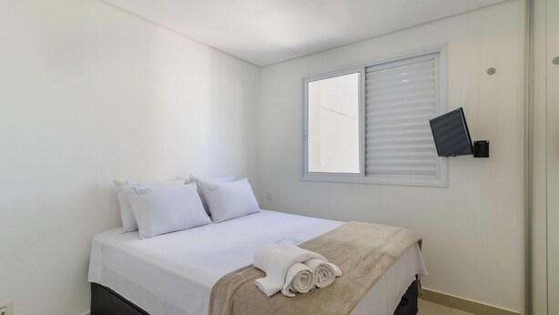 Quarto individual com uma cama branca, travesseiros brancos, um cobertor bege e uma TV fixada na parede.