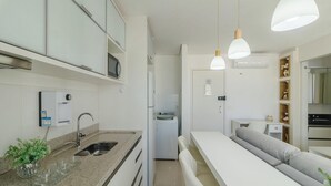 Uma cozinha compacta com micro-ondas integrado, pia e frigorífico. Possui uma área de refeições com uma mesa branca e um sofá cinzento.
