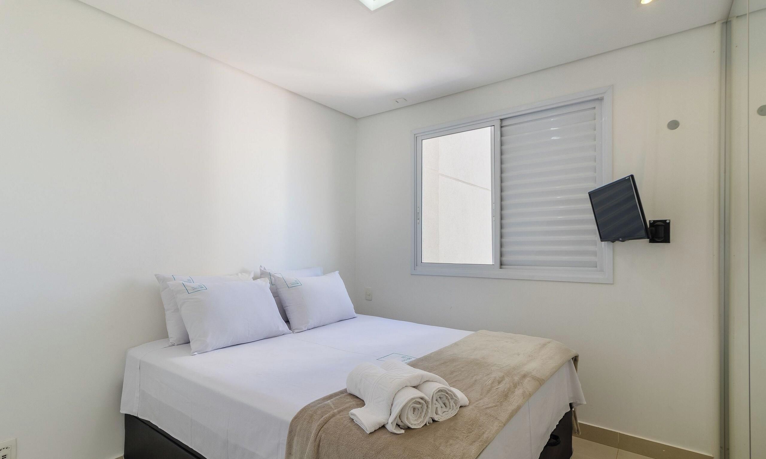 Une chambre à coucher avec un lit, des oreillers, une couverture, une fenêtre avec des stores et un téléviseur fixé au mur.