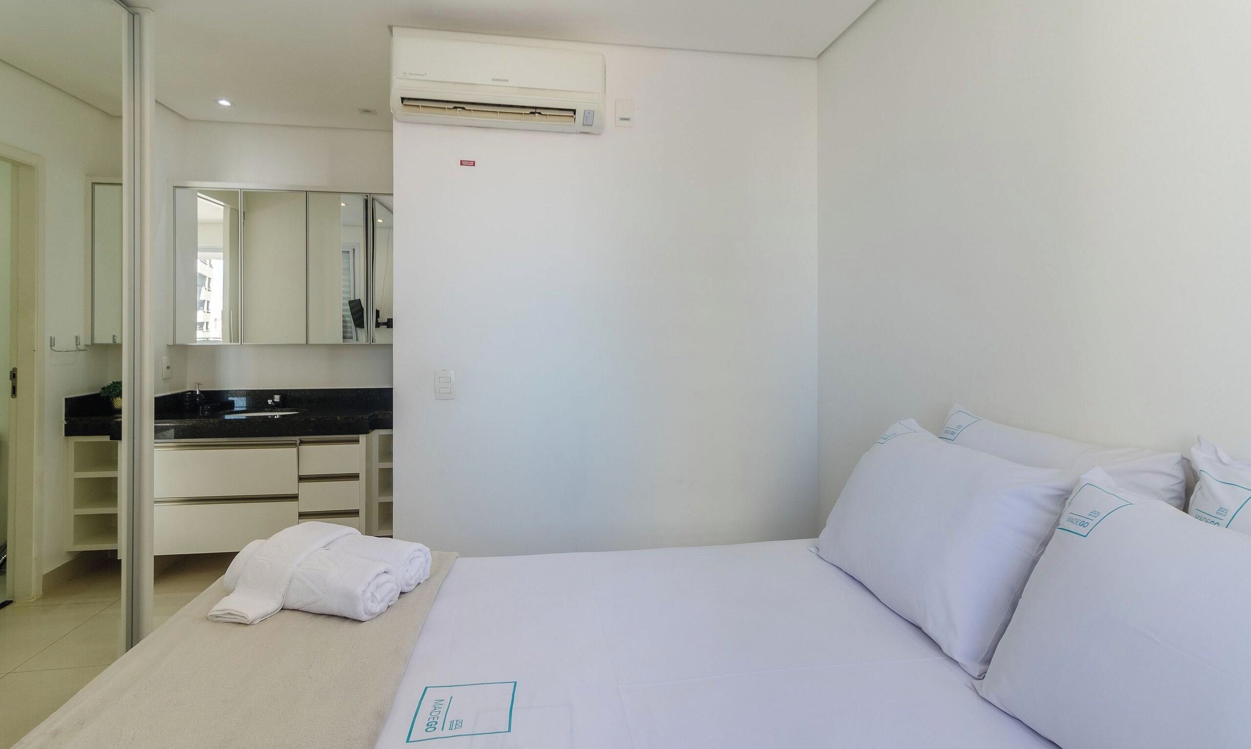 A hotel room with a bed, a bathroom with a sink and mirror, and an air conditioning unit.