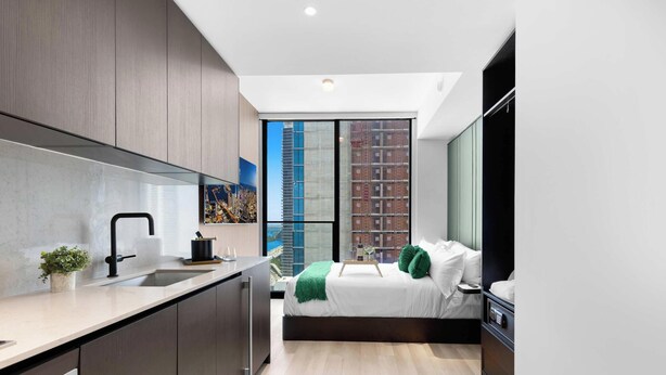 Quarto moderno com cama, mesa de cabeceira e vista para um edifício através de uma grande janela.