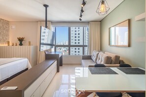 A modern hotel room with a bed, a sofa, a dining table, and a large window offering a city view. - Complete Studio in Condominium With Pool Dnag1702 (Goiânia)