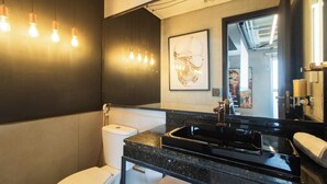 Ein modernes Badezimmer mit einem schwarzen Waschbecken, einer Toilette und einem Spiegel. Wandleuchten und ein gerahmtes Kunstwerk befinden sich über dem Spiegel.