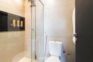 Ein modernes Badezimmer mit einer gläsernen Duschkabine, einer Toilette und einem schwarzen Ablageschrank mit Pflegeprodukten.