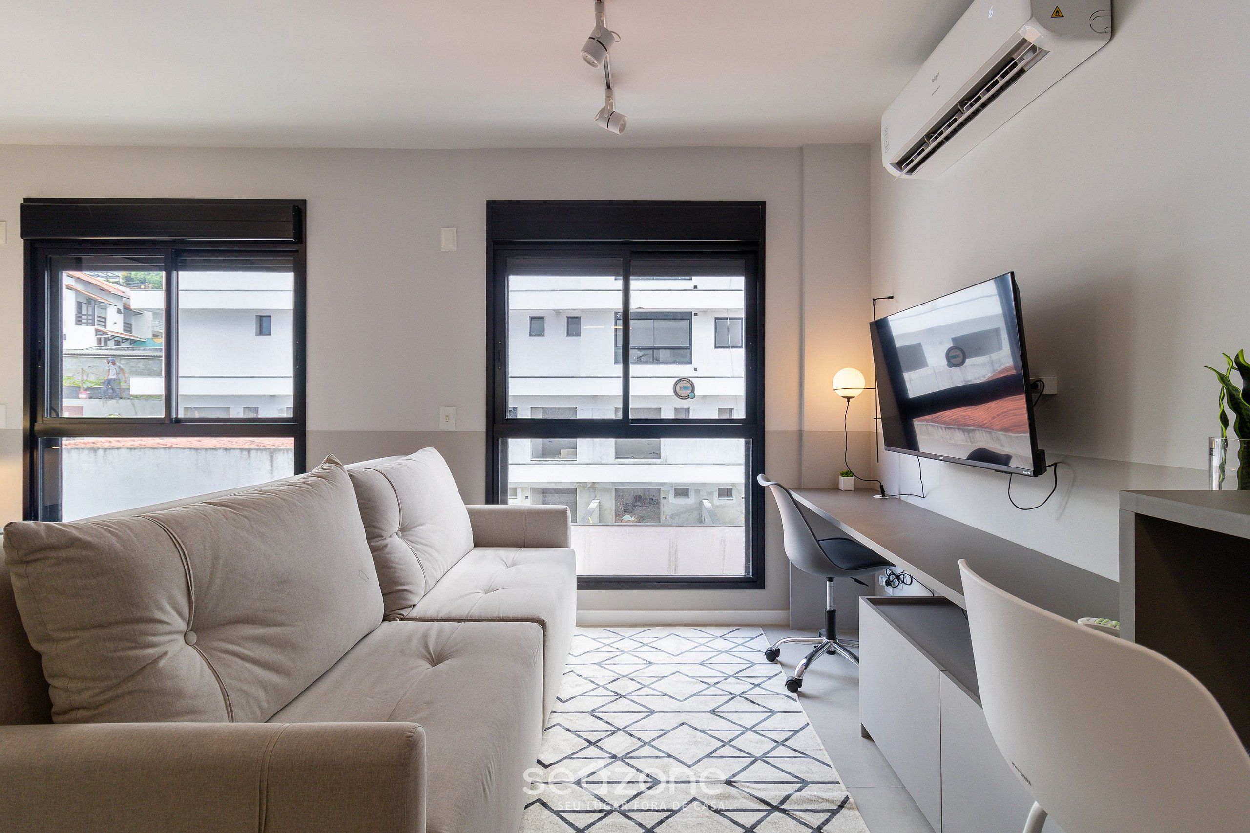 A modern living room with a sofa, a desk, and a TV. There are large windows and an air conditioning unit on the wall.