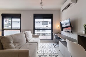Uma sala de estar moderna com sofá, escrivaninha e TV. Há janelas amplas e um aparelho de ar condicionado na parede.