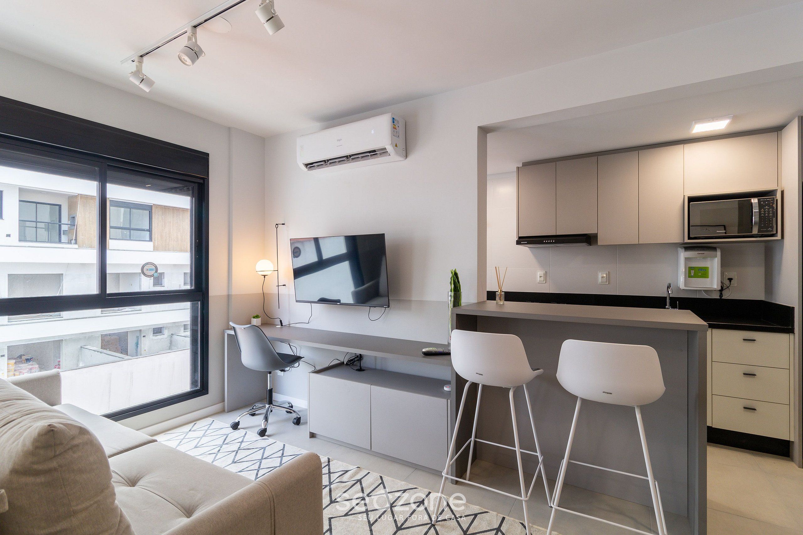 A modern kitchen with a flat-screen TV, a microwave, and a coffee machine. There is a bar area with stools and a large window providing a view of the city.