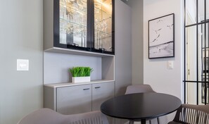 Um interior moderno com uma mesa redonda e duas cadeiras, um expositor de vidro com copos de vinho, uma obra de arte emoldurada e um vaso de plantas.