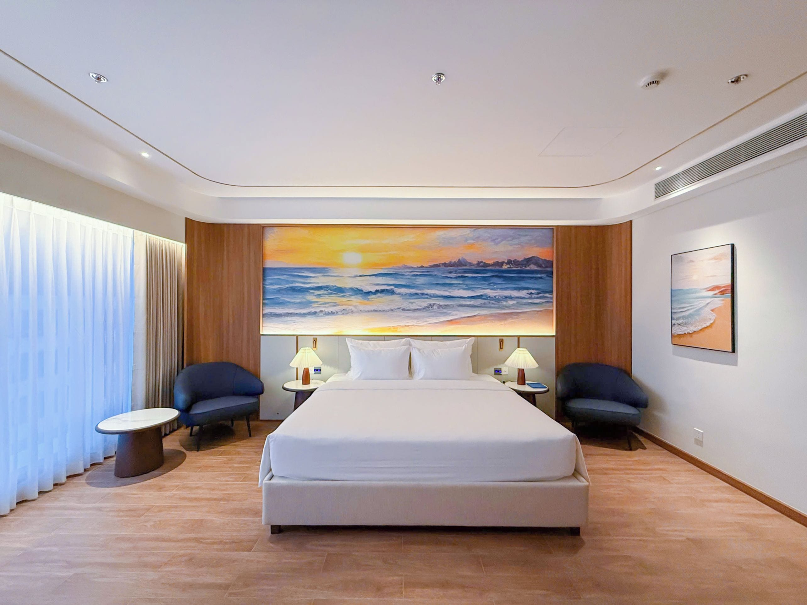 A hotel room with a large bed, two bedside tables, a nightstand, a chair, and a painting of a sunset over the ocean.