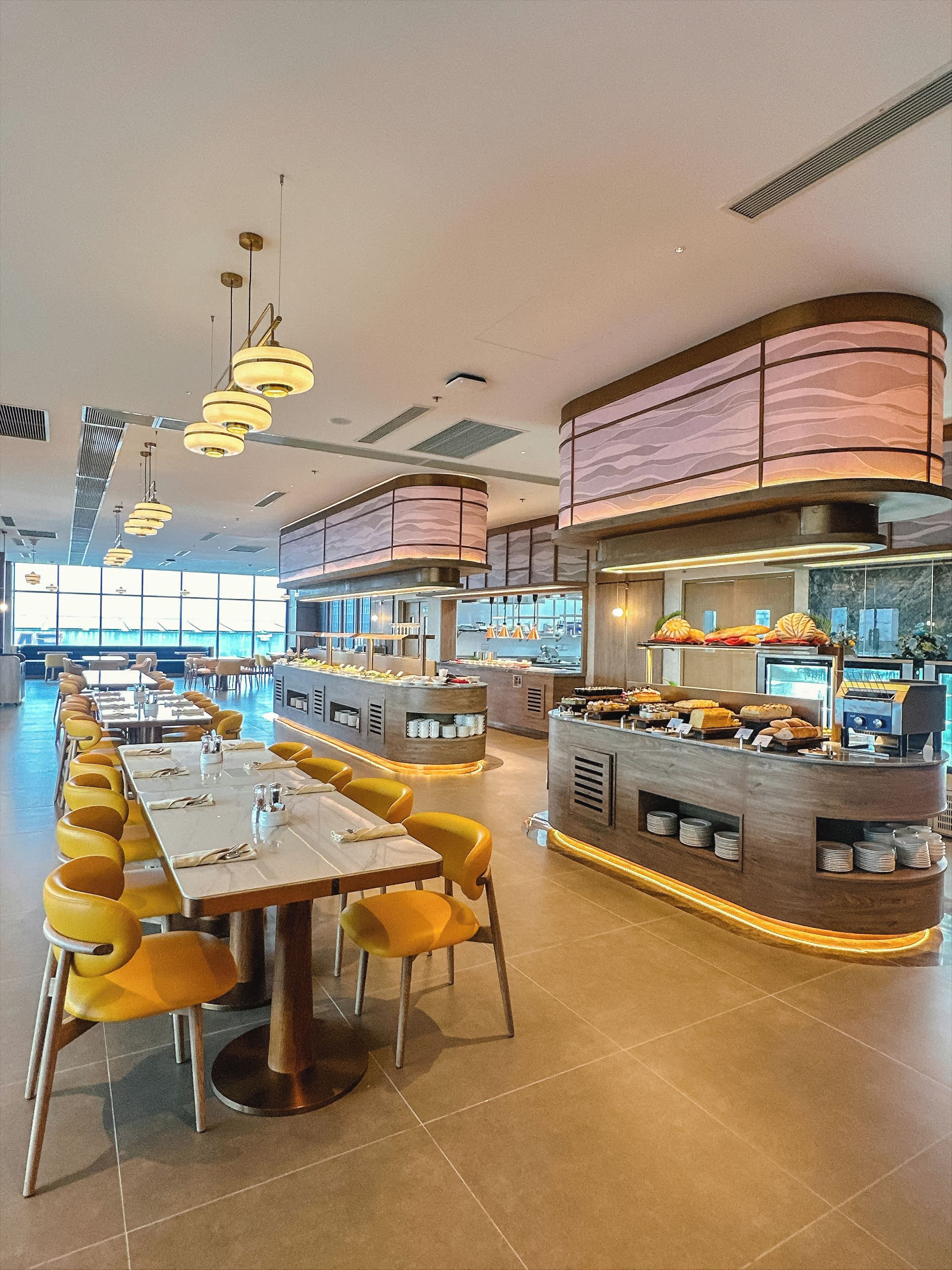 A modern restaurant with long communal tables and yellow chairs, a central buffet area, and a curved ceiling with patterned panels.