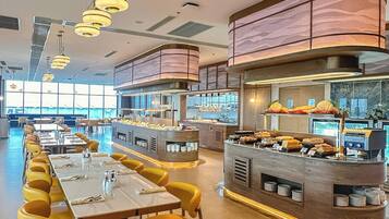 A modern restaurant with long communal tables and yellow chairs, a central buffet area, and a curved ceiling with patterned panels.