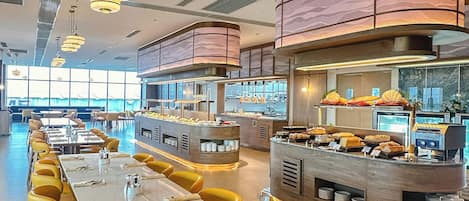 A modern restaurant with long communal tables and yellow chairs, a central buffet area, and a curved ceiling with patterned panels.