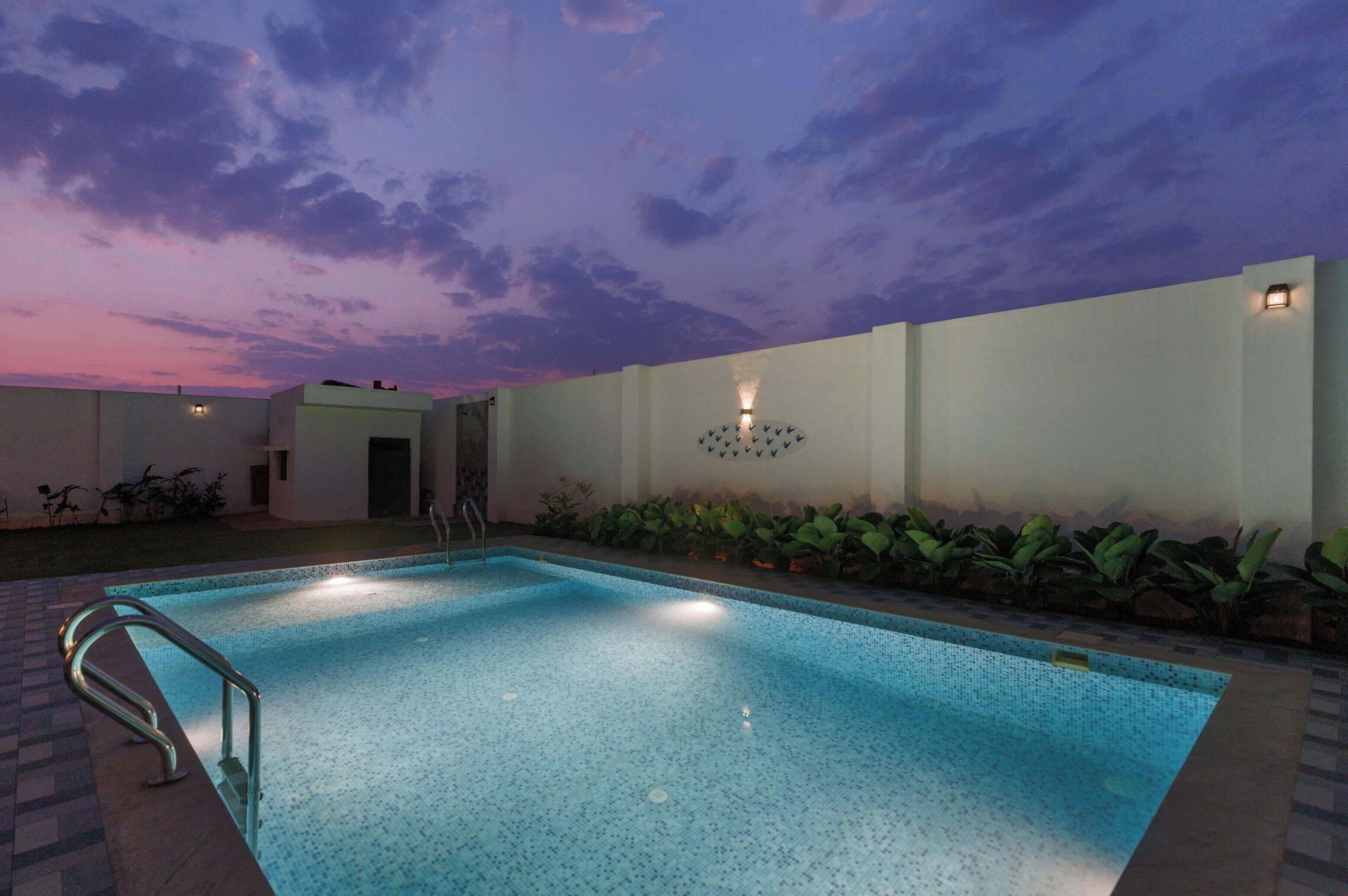 A swimming pool with a metal ladder, surrounded by a low wall with plants and a wall-mounted light fixture.