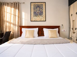 A hotel room with a large bed, a headboard with intricate woodwork, a framed artwork above the bed, a chair, and a wardrobe with a decorative pattern.