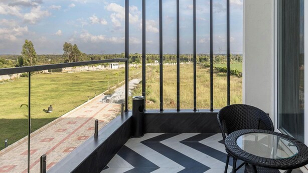 A balcony with a glass table and chairs, a chevron-patterned floor, and a view of a grassy field. - Crystal Blue Villa by Stayjadeprivatepoolvegfood (Doddaballapur)