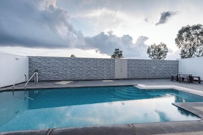 Una piscina con un muro de piedra, una escalera metálica y una mesa con sillas.