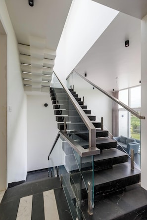 A modern staircase with glass railings and a view of the outdoors. - Crystal Blue Villa by Stayjadeprivatepoolvegfood (Doddaballapur)