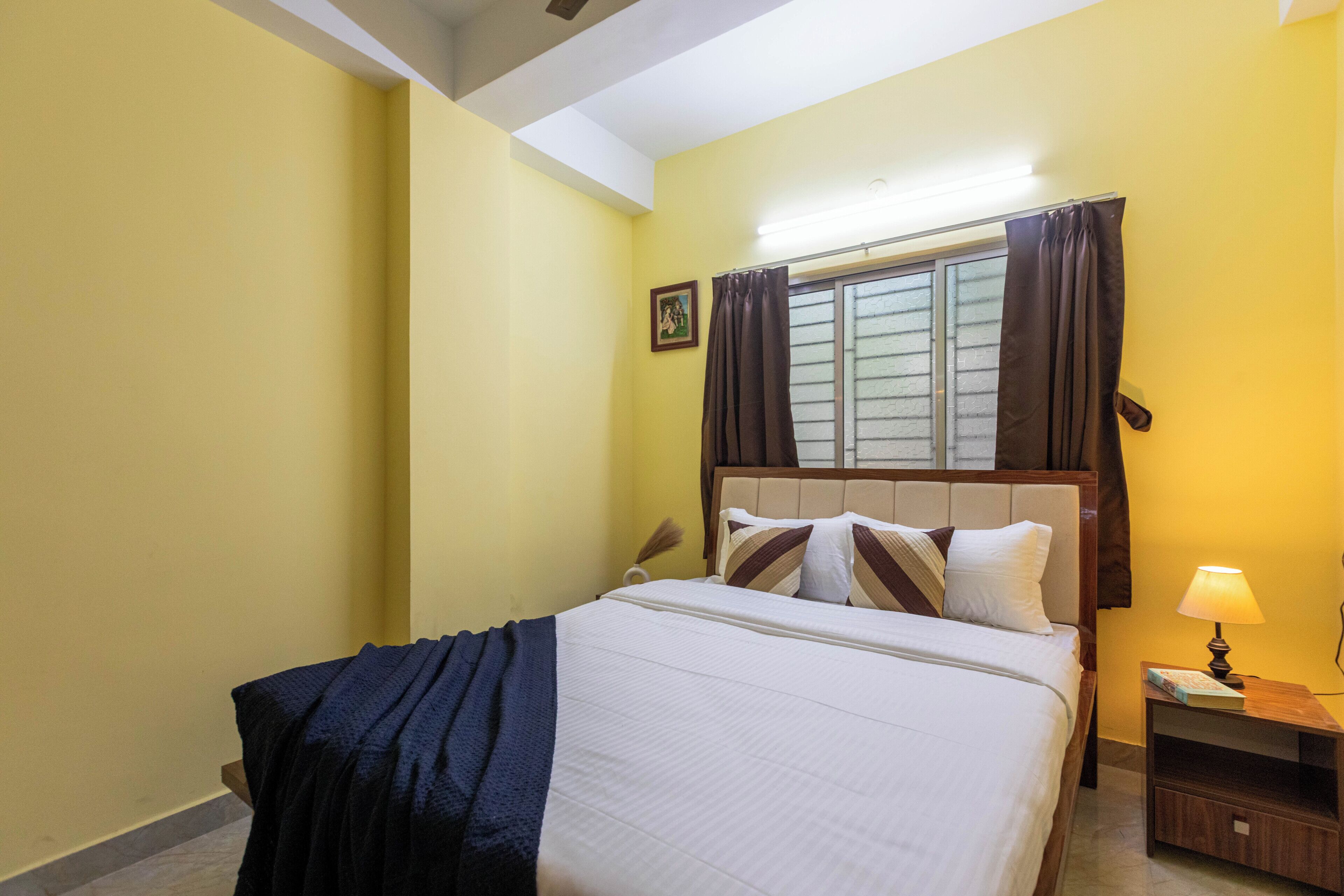 A bedroom with a bed, a nightstand, a lamp, a window with curtains, and a picture on the wall.