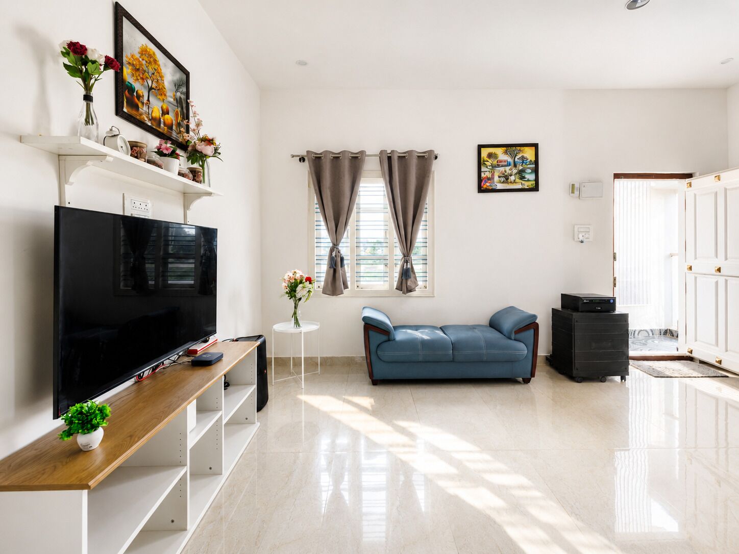 A modern living room with a flat-screen TV, a blue sofa, a small white table with a vase, and a window with curtains.