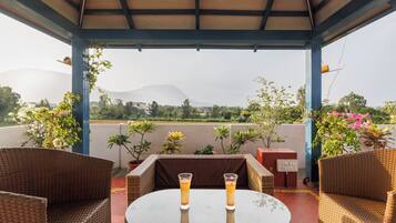 A covered outdoor seating area with wicker furniture and two glasses of a beverage on a table, overlooking a landscape with hills.