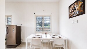 Uma sala de jantar com uma mesa branca posta para quatro pessoas, uma geladeira e uma janela com persianas.