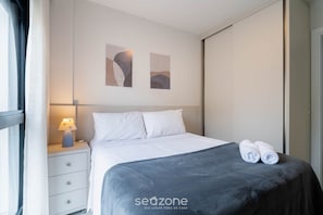 A neatly made bed with white linens, a grey blanket, and two rolled towels on the foot of the bed. There is a bedside table with a lamp and a wall-mounted artwork above it. - Complete Studio in Floripa Downtown Ech0205 (Florianópolis)