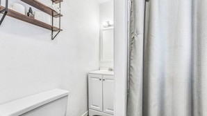 A bathroom with a white toilet, a white sink, and a mirror above it. There is a shower curtain visible on the right side. - Coffield - Comfort, Privacy, and Convenience in Killeen, Texas (Killeen)