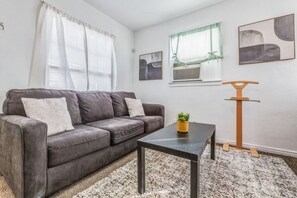 Uma sala de estar com um sofá cinzento, uma mesa de centro, uma planta pequena e um arranhador de madeira para gatos.