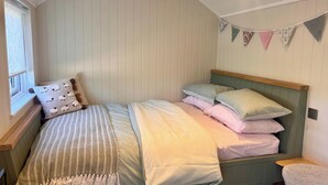 A bedroom with a bed, pillows, a window with curtains, and a wall with bunting. - Peaceful Shepherd Hut Near Clovelly (Bideford)