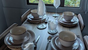 A dining table set with plates, bowls, glasses, and cutlery, with a view of greenery through a window.