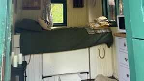 A small, cozy room with a single bed, a window with curtains, a small table with candles, and a dresser. - Fern Cosy Woodland Shepherds Hut Walk to Sea (Holyhead)
