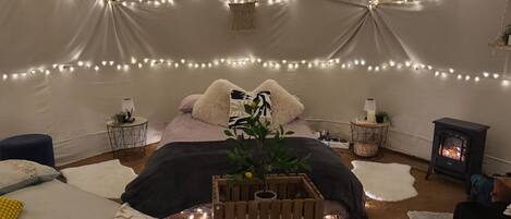 A cozy tent-style room with a bed, bedside tables, a small heater, string lights, and a potted plant.