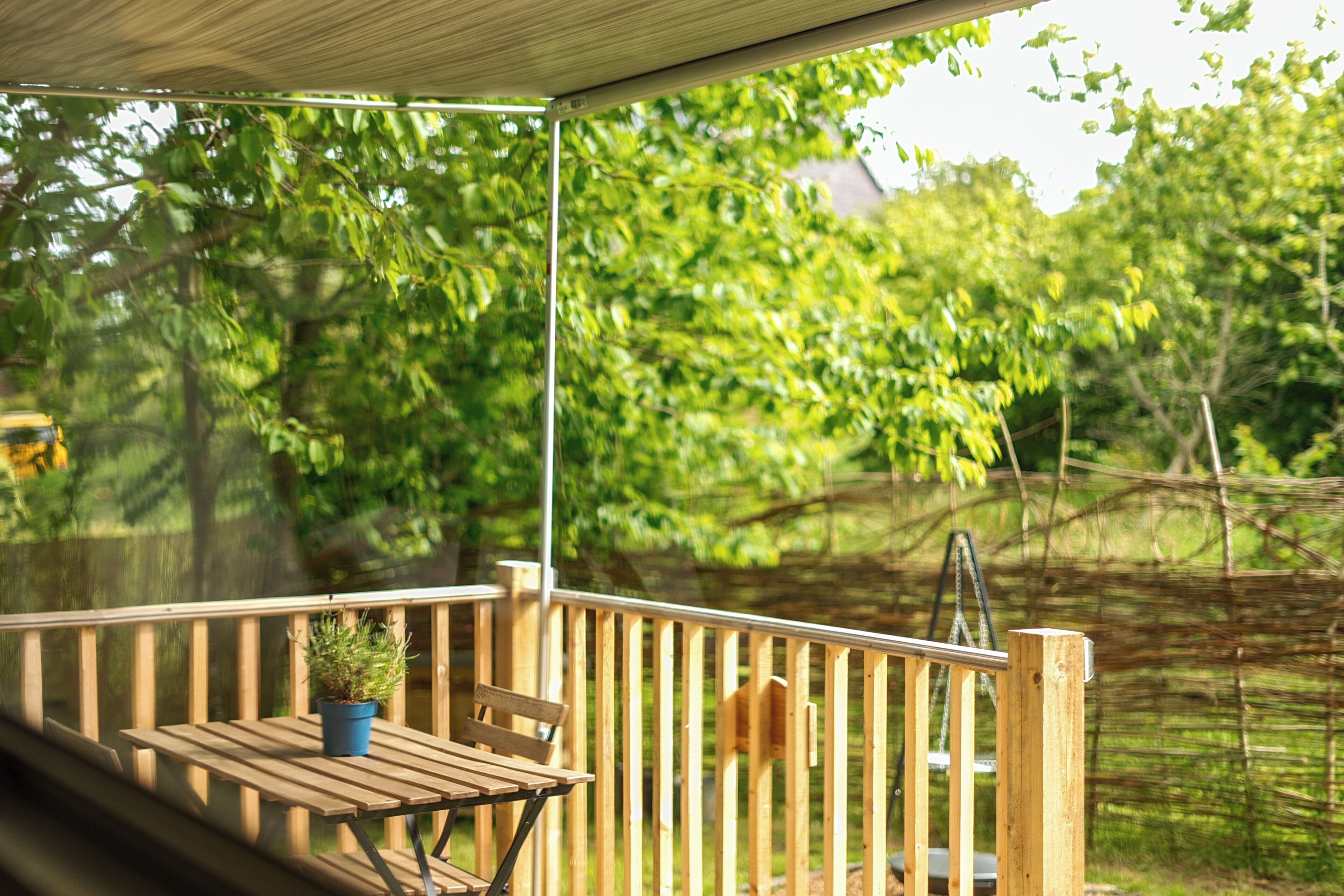 A wooden deck with a table and chair, a potted plant, and a view of a green, leafy area.