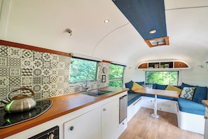 A compact kitchen with a sink, stove, and a kettle. There is a seating area with cushions and a window overlooking greenery. - Sleeps 2 - Private Hot Tub - Fire Pit (Cardigan)