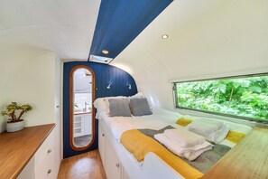 Quarto compacto com cama, mesa de cabeceira, espelho e janela com vista para vegetação.