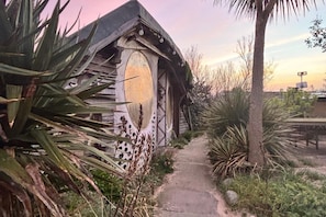 Un edificio rústico con un diseño arquitectónico singular, un sendero que lo lleva a la entrada y vegetación circundante.