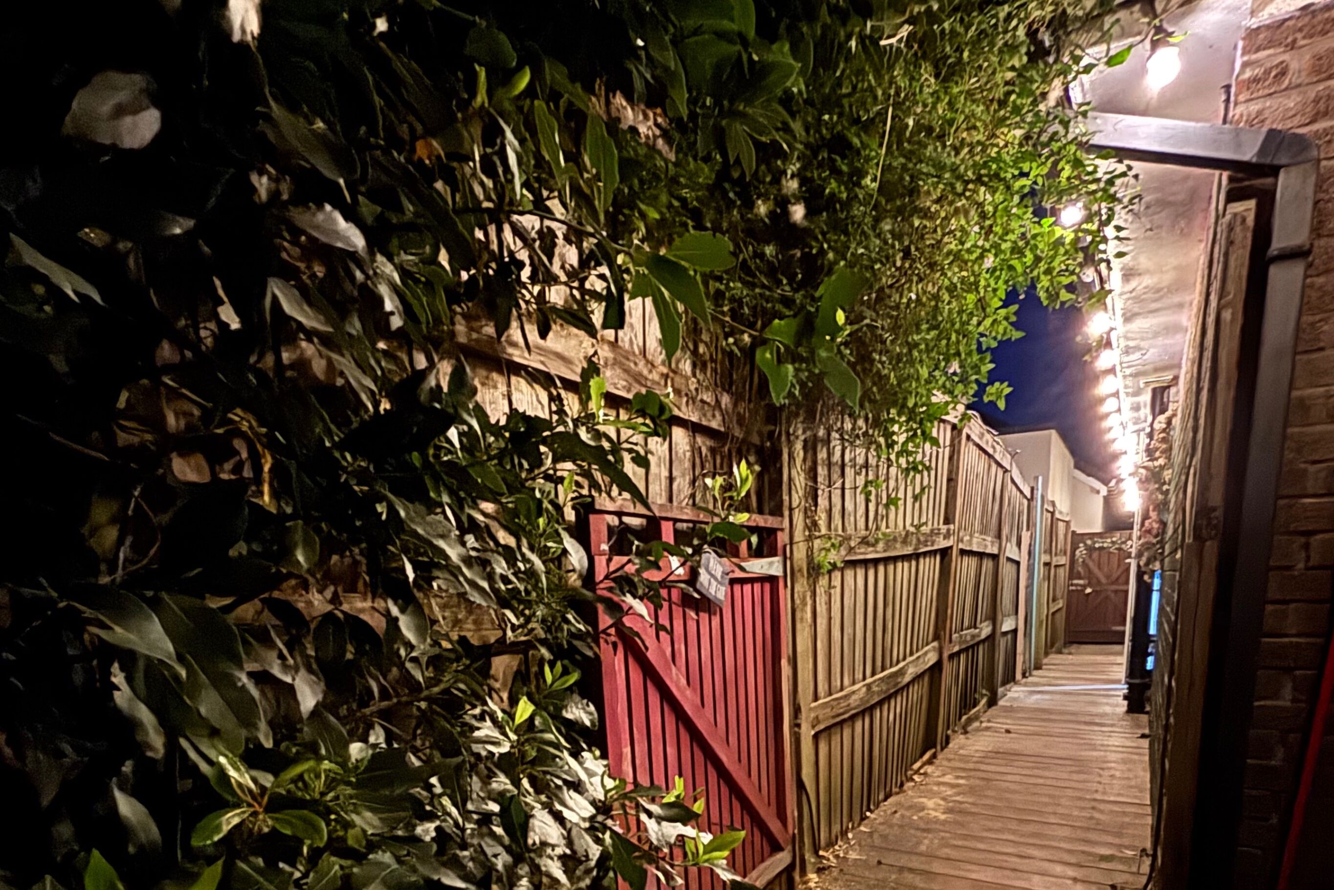 A narrow wooden pathway flanked by dense foliage and a red gate, leading to a building with a stone facade.