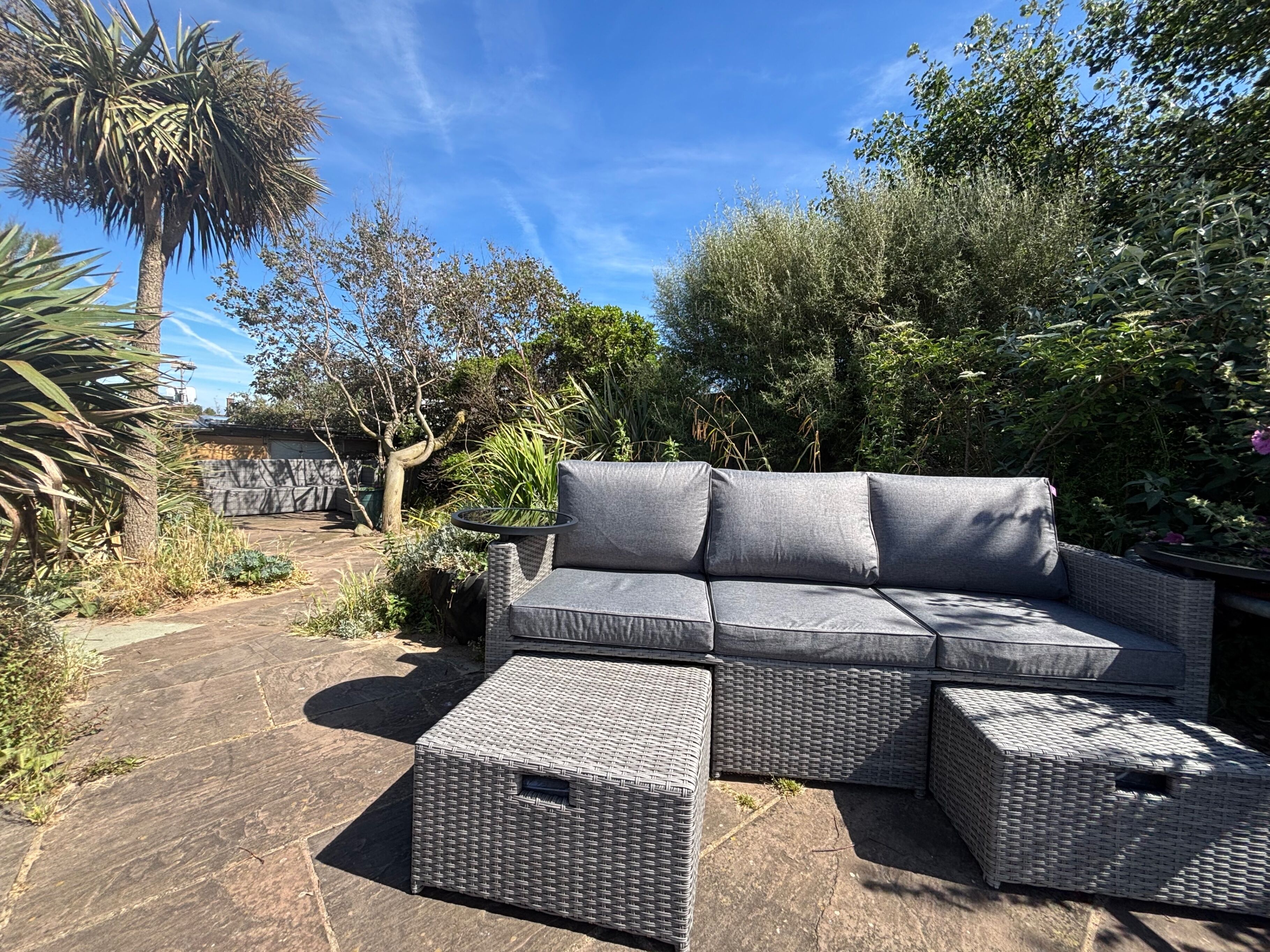 A patio area with a grey wicker sofa and ottomans, surrounded by lush greenery and a clear blue sky.