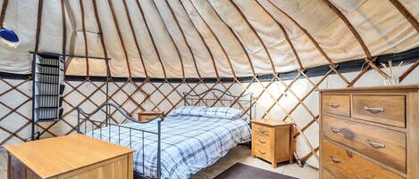 A cozy interior of a round tent with a bed, wooden dresser, and a small shelf.