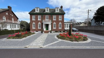 A two-story brick building with a green door, a driveway with gravel, and red flowers in the foreground.