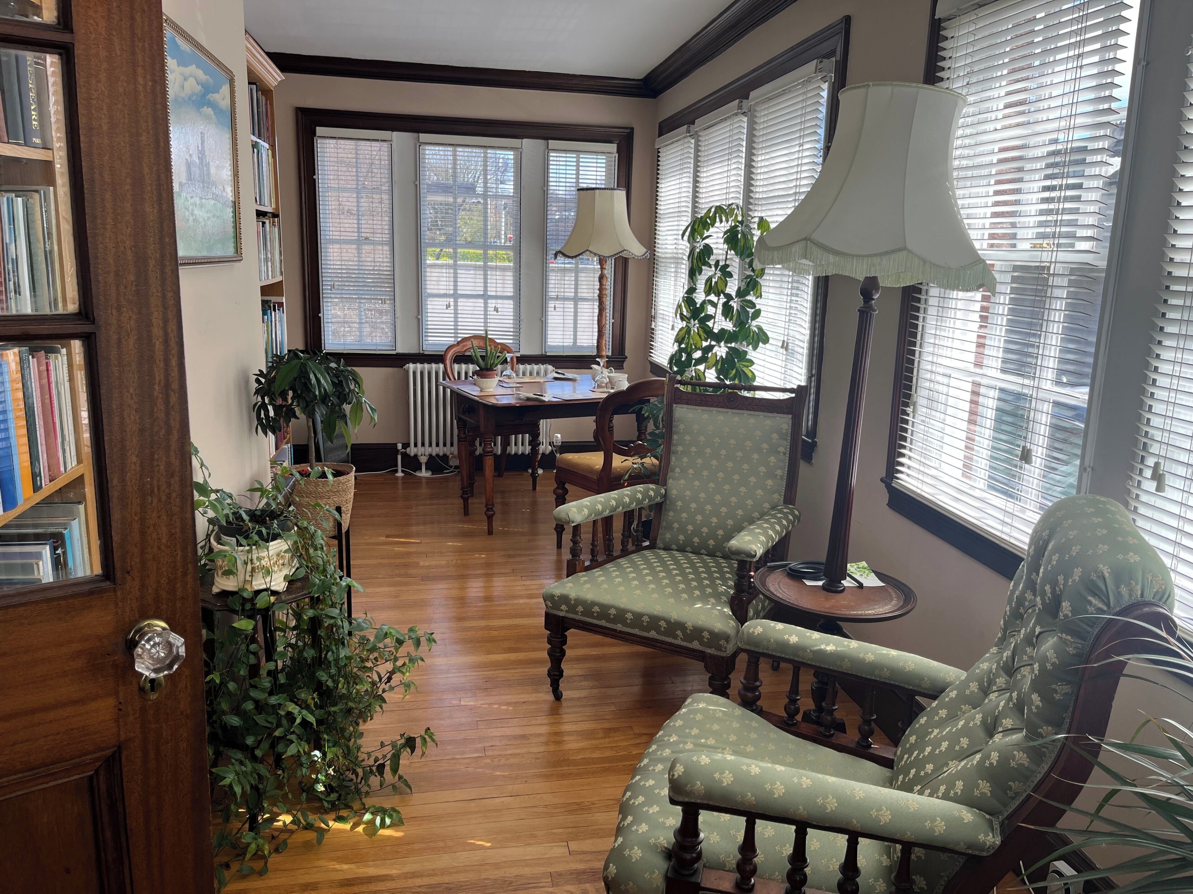 A room with a wooden floor, a dining table with chairs, a bookshelf, and a large window with blinds.