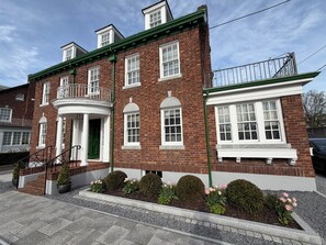 A two-story brick building with a green front porch, white window frames, and a small balcony with a metal railing.