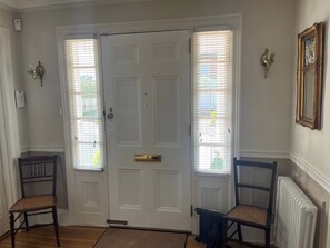 A room with a white door, two windows with blinds, a radiator, and two chairs.