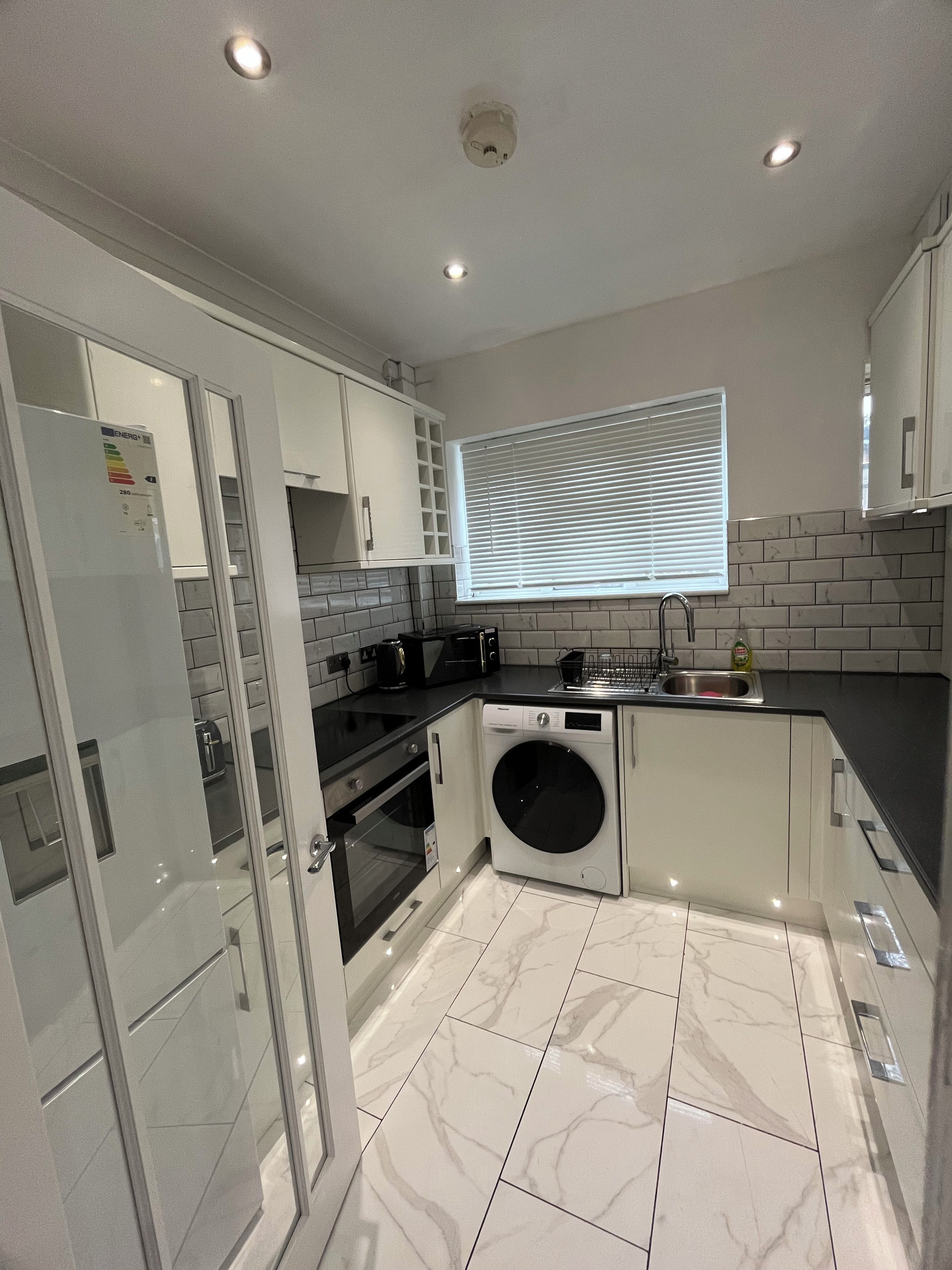 A modern kitchen with a built-in oven, microwave, and washing machine, a window with blinds, and a tiled backsplash.