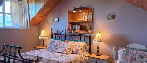 A cozy bedroom with a sloped ceiling, a bed with floral bedding, two bedside tables with lamps, and a bookshelf.